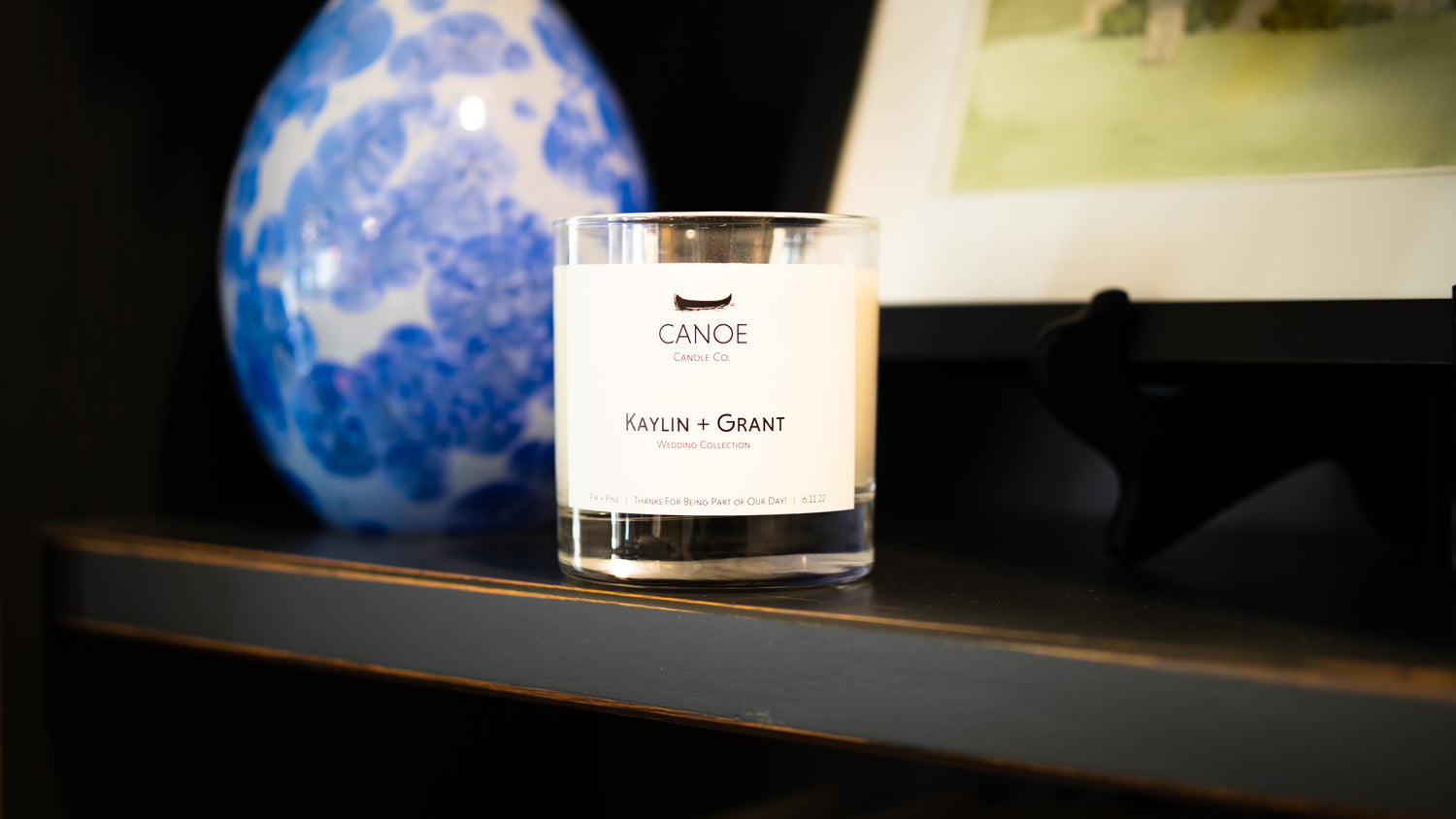 Canoe Candle Co's Canoe Custom 10oz soy wax candle sitting on a black wooden shelf in front of a blue pottery vase and framed water-colour print.