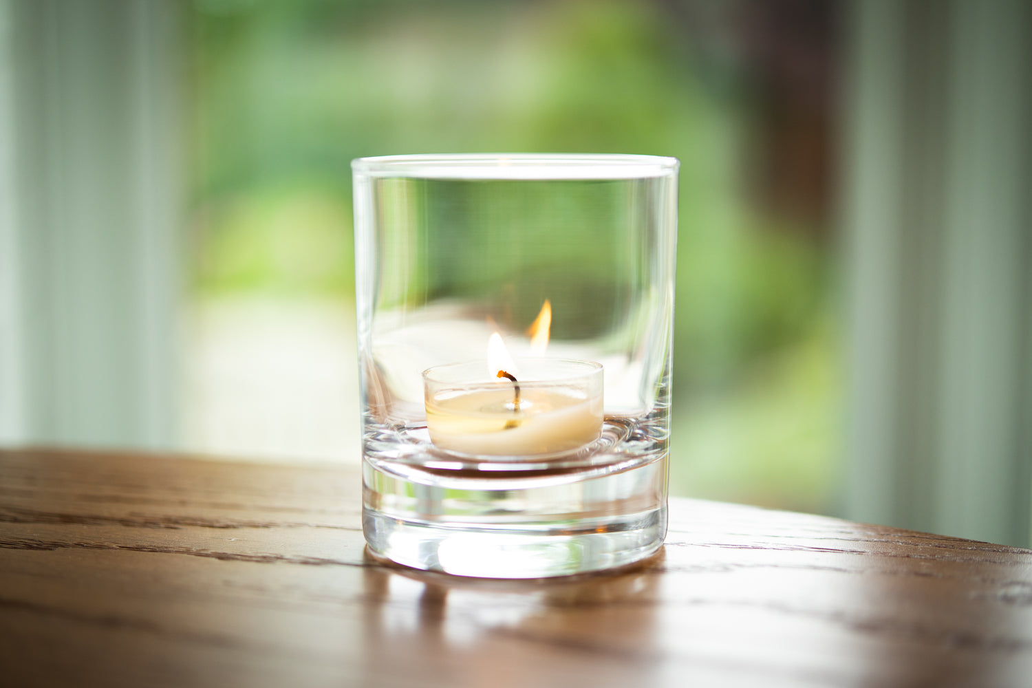 Canoe Candle Co's 7oz tealight rocks glass with tealight burning inside, sitting on a wooden table in front of window.