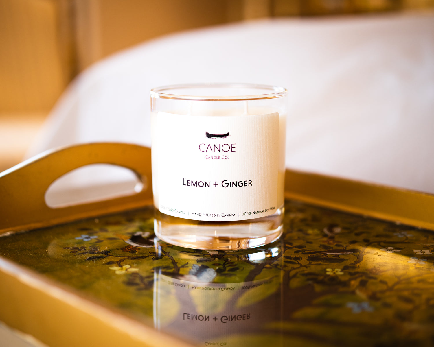 CANOE Candle Co.'s Lemon + Ginger 10oz soy wax candle on a tray in a bedroom with white linen sheets in the background.