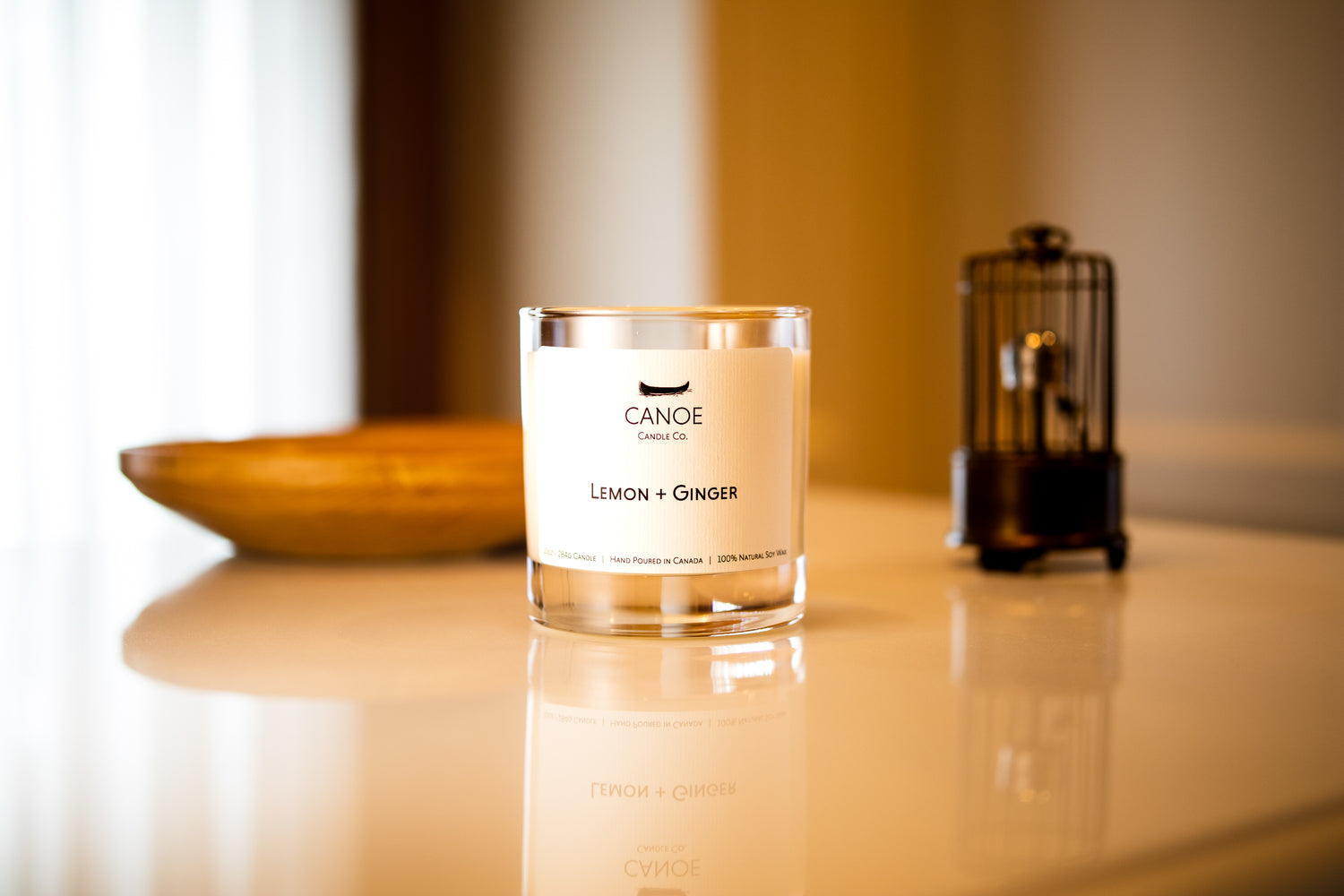 Canoe Candle Co.’s Lemon + Ginger 10oz soy wax candle on a white marble counter in front of a window and closet.