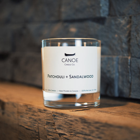Canoe Candle Co.’s Neroli + Driftwood 10oz soy wax candle on a rough sawn mantle in front of dry stacked grey stone.