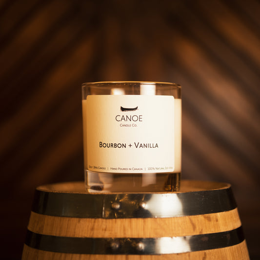 Canoe Candle Co.’s Bourbon + Vanilla 10oz soy wax candle on whisky barrel with dark wood background.