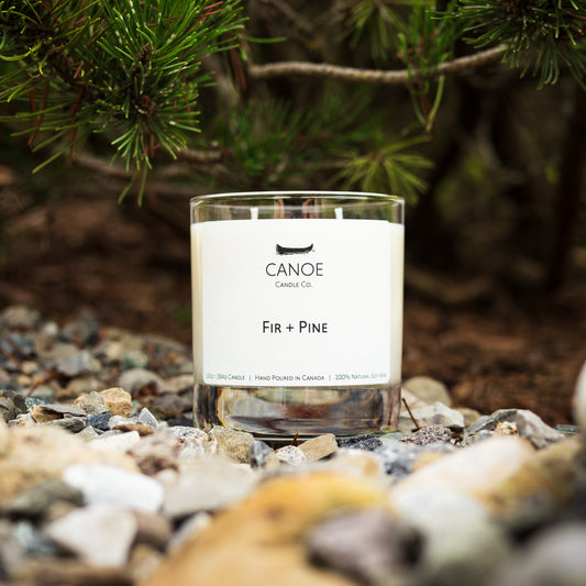 Canoe Candle Co.’s Fir + Pine 10oz soy wax candle on stone with pine branches overhead.