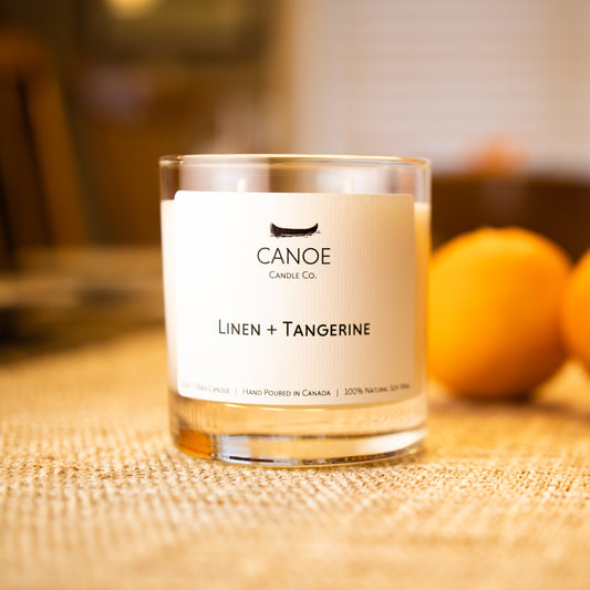 Canoe Candle Co.’s Linen + Tangerine 10oz soy wax candle on linen table runner in front or two oranges.