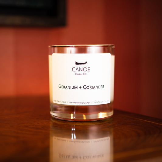 Canoe Candle Co.’s Geranium + Coriander 10oz soy wax candle on dark wood table with red wallpaper in the background.