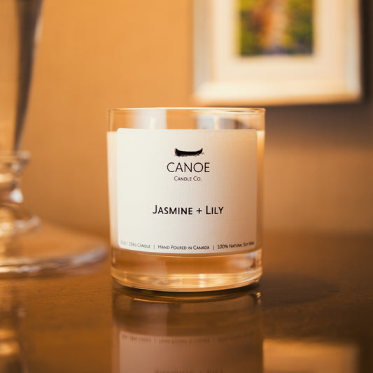 Canoe Candle Co.’s Jasmine + Lily 10oz soy wax candle on dark wood table with glass lamp and small painting in the background.