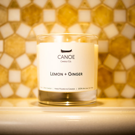 Canoe Candle Co.’s Lemon + Ginger 10oz soy wax candle on marble shelf in front of white and yellow patterned marble backsplash.